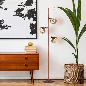 "A modern floor lamp with three adjustable shades in a wood-like and brass finish stands next to a wooden dresser, artwork, and a plant in a woven basket."