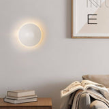 Rochi Wall Lamp in white aluminum mounted in a modern living room, casting a soft glow above a wooden side table with books.