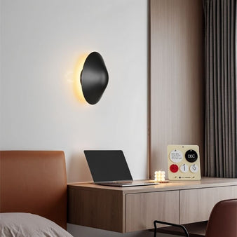 Rochi Wall Lamp in black aluminum mounted above a modern desk setup, casting warm ambient light in a contemporary bedroom.