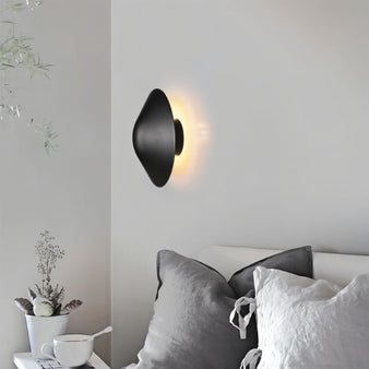 Rochi Wall Lamp in black aluminum illuminating a cozy bedroom corner with soft ambient lighting, complementing neutral decor.