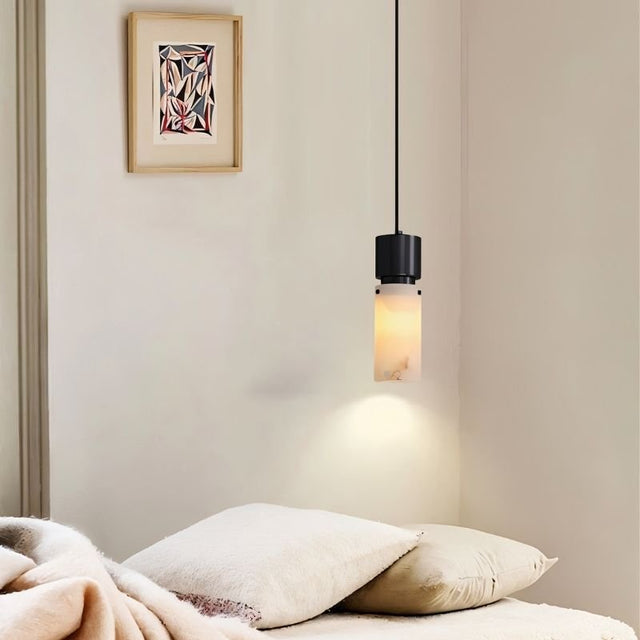 "Modern pendant light with a cylindrical alabaster shade and a black frame, illuminating a small space in a bedroom."
