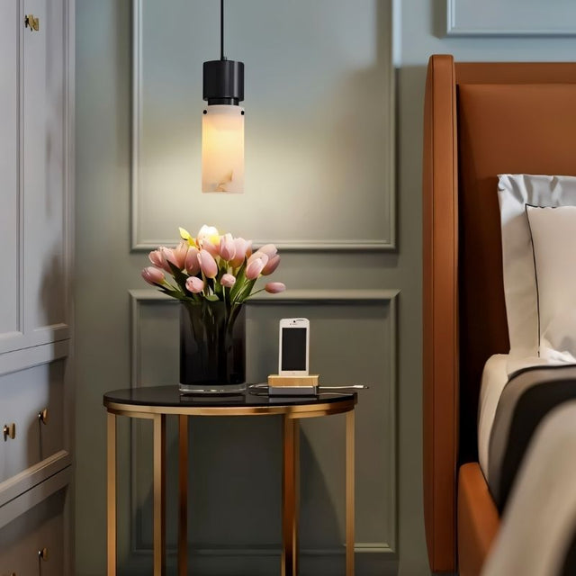 "Modern pendant light with a cylindrical alabaster shade and a black frame, illuminating a bedside table in a contemporary bedroom."