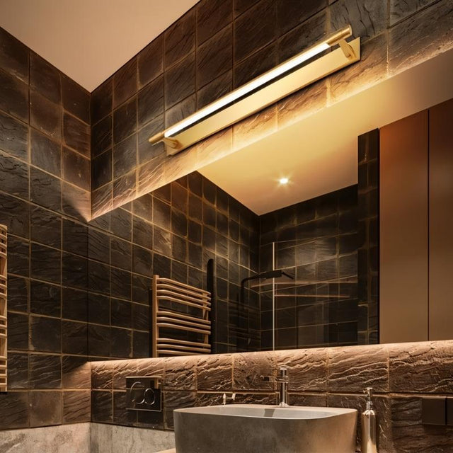 "A modern gold wall lamp with a sleek, adjustable cylindrical light bar mounted above a large mirror and sink in a contemporary bathroom with dark tiles."