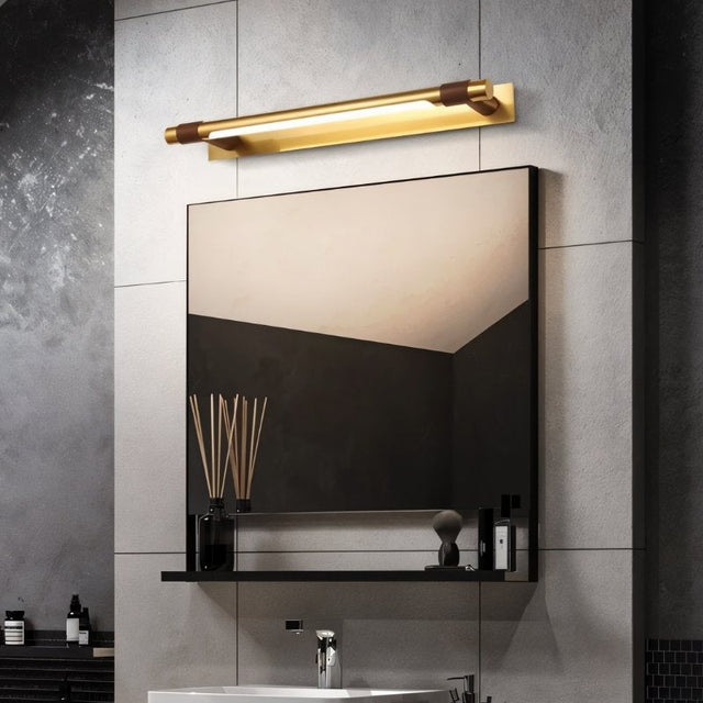 "A modern gold and brown wall lamp with a sleek, adjustable cylindrical light bar mounted above a rectangular mirror in a contemporary bathroom with a minimalist shelf."