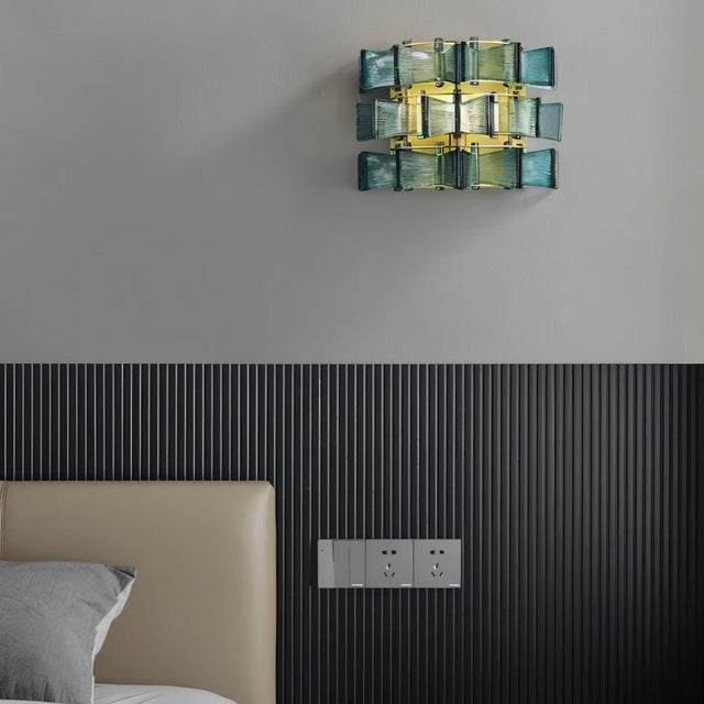 "Iron Salane wall lamp with layered blue glass panels, mounted on a grey wall above a bed with a beige headboard."