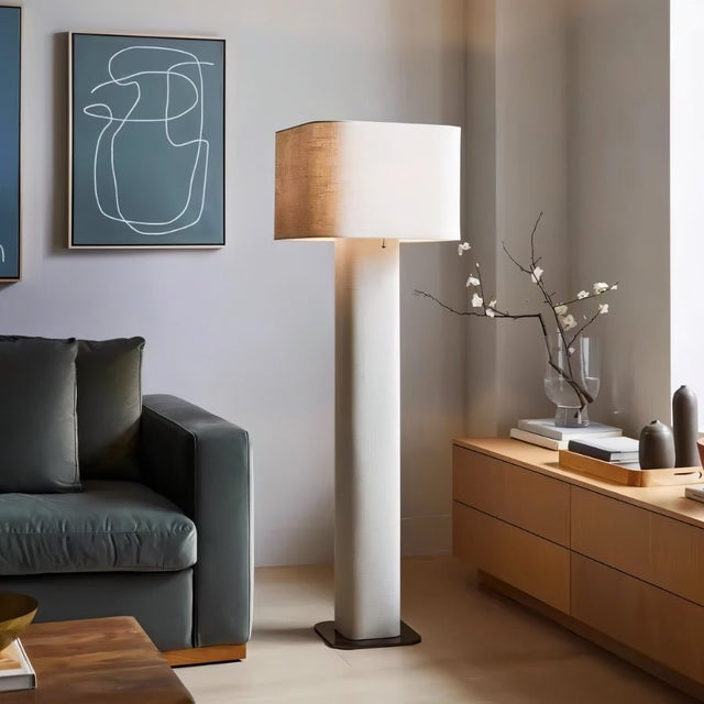 "Modern floor lamp with a cylindrical iron base and a fabric shade in a contemporary living room setting with a sofa and artwork."