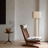 "Modern floor lamp with a cylindrical iron base and a fabric shade illuminating a reading nook with a wooden chair, a side table, and a book."
