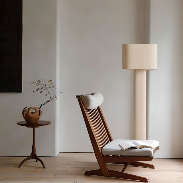 "Modern floor lamp with a cylindrical iron base and a fabric shade illuminating a reading nook with a wooden chair, a side table, and a book."