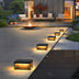 Black recessed outdoor garden lamps with warm LED lighting along a modern patio pathway at dusk.