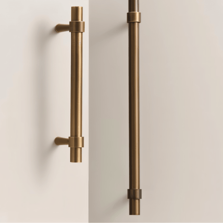 "Two antique brass pull bars with cylindrical design and subtle accents, mounted vertically on white cabinet doors."