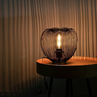 "A small, rounded black table lamp with an open-wire metal frame, casting a warm glow on a round wooden side table in a dimly lit bedroom."