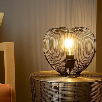 "A small, rounded black table lamp with an open-wire metal frame, radiating a warm light on a wire-mesh side table, creating an industrial aesthetic."