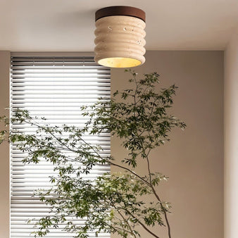 "A retro-inspired ceiling light with a travertine shade and a wooden base, mounted on a living room ceiling, providing a warm, ambient glow near a window with blinds and a plant."