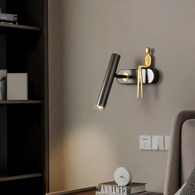 "A black and gold bedside reading lamp with a figurine on a wall next to a bed."