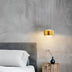 Gold cylindrical pendant light hanging above a bedside table in a modern bedroom with concrete walls.