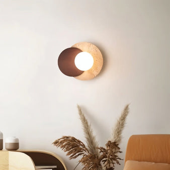 "A wabi-sabi wall lamp with travertine and wood elements, mounted on a light-colored living room wall, providing a soft, ambient glow above a modern console table and dried pampas grass."