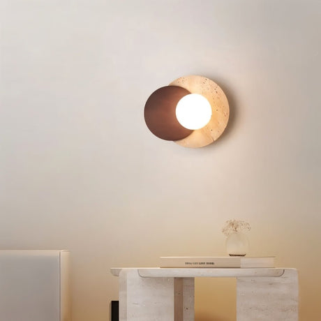 "A wabi-sabi wall lamp with travertine and wood elements, mounted on a light-colored wall in a modern home, providing a soft, ambient glow above a minimalist table with a book and a vase."