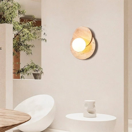 "A wabi-sabi wall lamp featuring travertine and wood, mounted on a light-colored wall in a modern living room, providing a soft, ambient glow near a round table and a white armchair."