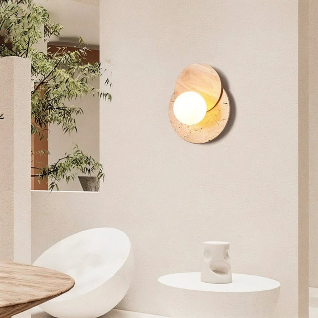 "A wabi-sabi wall lamp featuring travertine and wood, mounted on a light-colored wall in a modern living room, providing a soft, ambient glow near a round table and a white armchair."