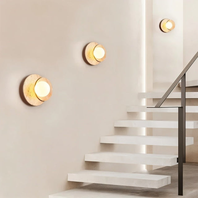 "Three wabi-sabi wall lamps with travertine and wood elements, arranged on a wall alongside a modern staircase, providing a soft, ambient glow."