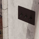 Three-gang matte black The Brass Toggle Switch (1 to 5 Gang) - Open Box installed on rustic plaster wall, blending vintage and modern aesthetics.
