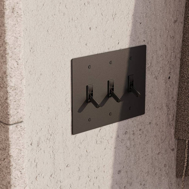 Triple gang The Brass Toggle Switch (1 to 5 Gang) - Open Box in matte black finish, mounted on textured concrete wall for modern industrial decor.