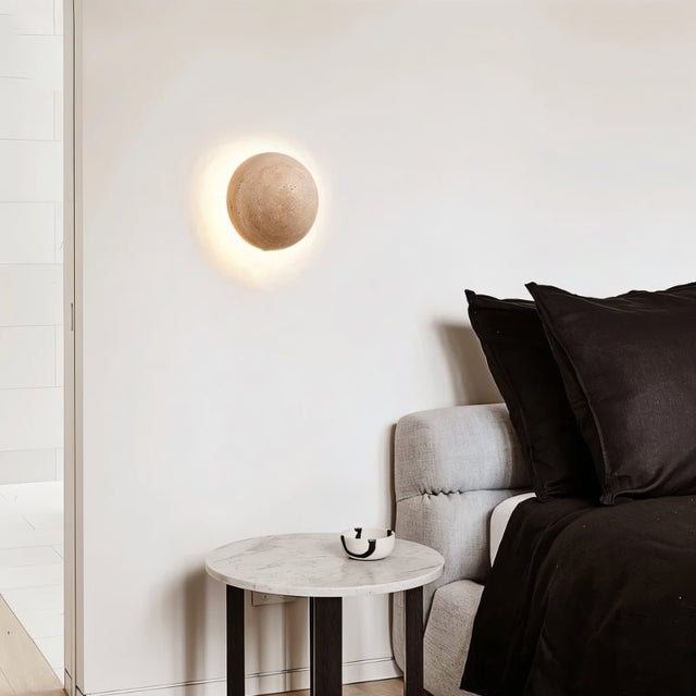 "A spherical travertine wall lamp emitting a soft, warm glow, mounted on a white wall in a modern bedroom above a side table and a bed with black linens."
