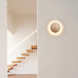 "A spherical travertine wall lamp emitting a warm glow, mounted on a white wall in a modern hallway with a wooden staircase."