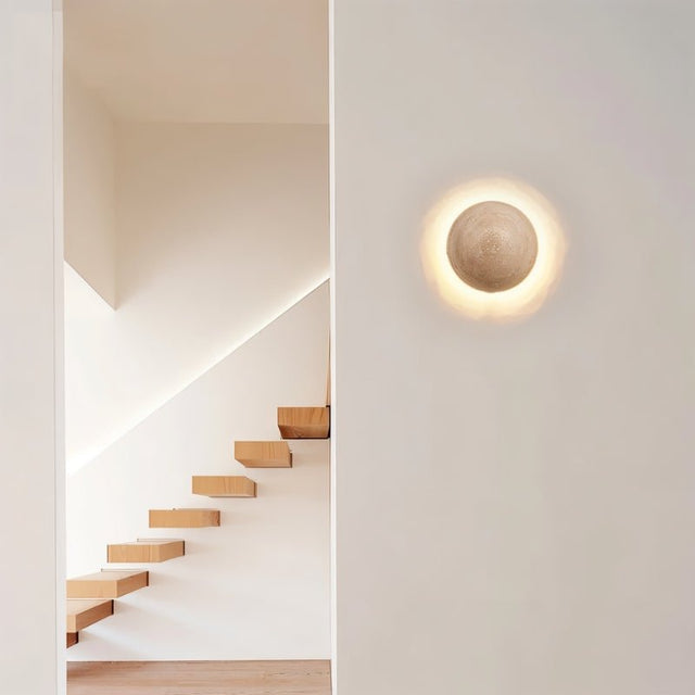 "A spherical travertine wall lamp emitting a warm glow, mounted on a white wall in a modern hallway with a wooden staircase."