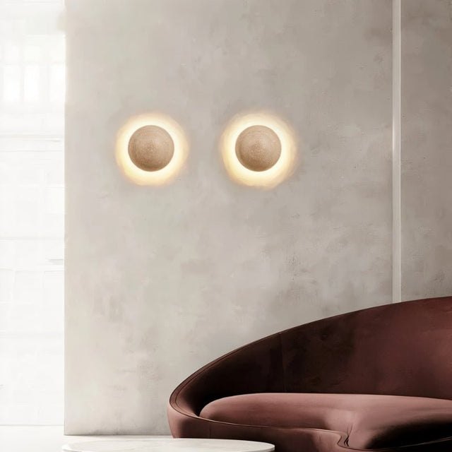 "Two spherical travertine wall lamps emitting a warm, radiant glow, mounted on a textured wall in a modern living room above a velvet sofa."