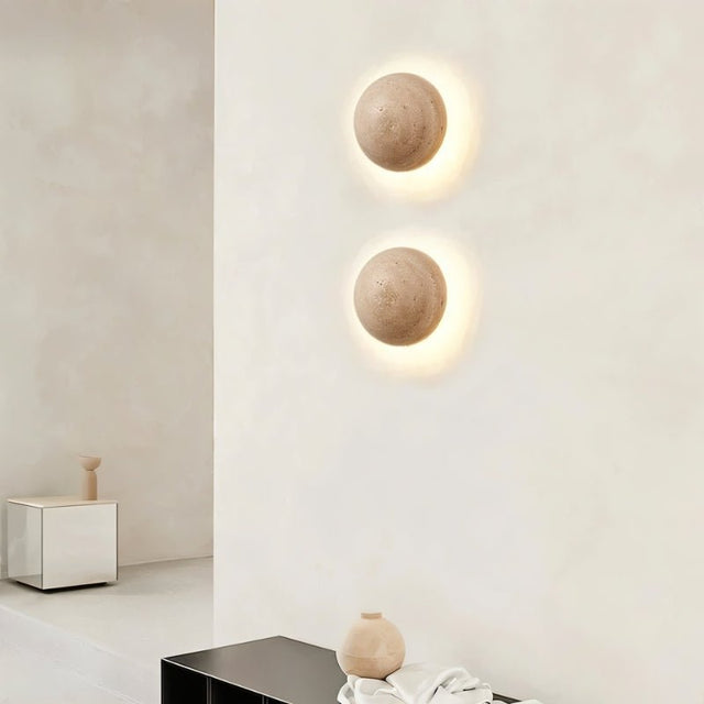 "Two spherical travertine wall lamps emitting a warm, radiant glow, mounted on a white wall in a modern space."