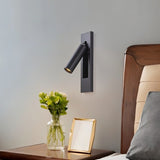 "A black wall lamp with a rectangular base and a cylindrical spotlight, shown mounted on a light gray wall above a nightstand with flowers in a clear vase and a gold picture frame, next to a bed with a gray pillow."