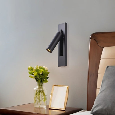 "A black wall lamp with a rectangular base and a cylindrical spotlight, shown mounted on a light gray wall above a nightstand with flowers in a clear vase and a gold picture frame, next to a bed with a gray pillow."