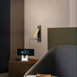 "A black and gold wall lamp with a rectangular base and a cylindrical spotlight, shown mounted on a light gray wall above a nightstand with a phone charging on a stand, next to a chair."