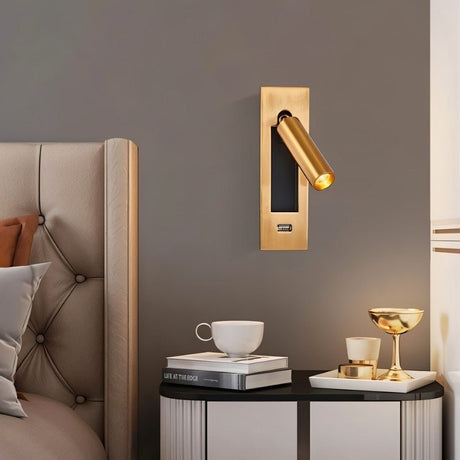 "A gold wall lamp with a rectangular base and a cylindrical spotlight, shown mounted on a gray wall above a nightstand with a book, cup, and gold decorative items, next to a bed with a tufted headboard."