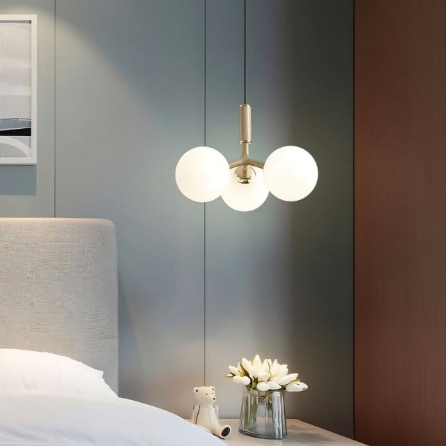 "A modern pendant light with three opal glass globes, in gold, hanging above a nightstand with flowers and a teddy bear."