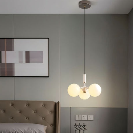 "A modern pendant light with three opal glass globes, in white and gold, hanging in a bedroom with a tufted headboard and artwork."