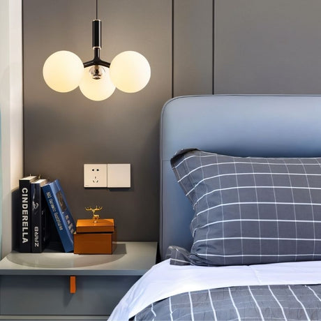 "A modern pendant light with three opal glass globes, in black, hanging above a nightstand with books and a decorative box in a bedroom."