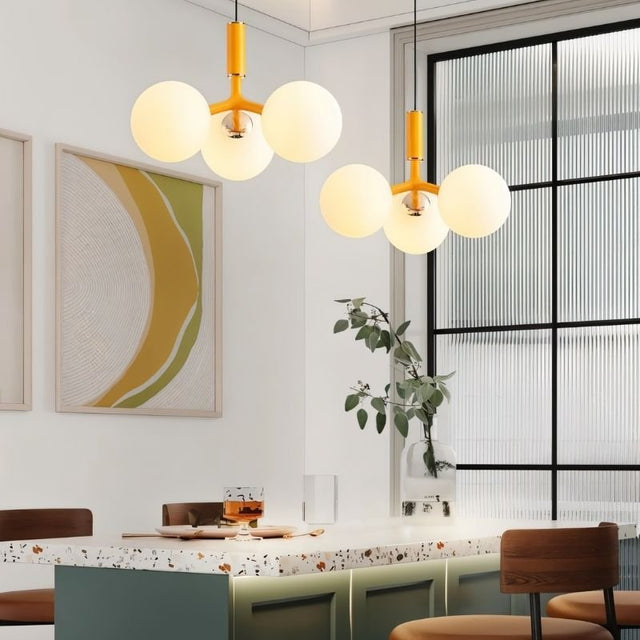 "Two modern pendant lights with three opal glass globes each, in orange, hanging over a dining table with chairs."