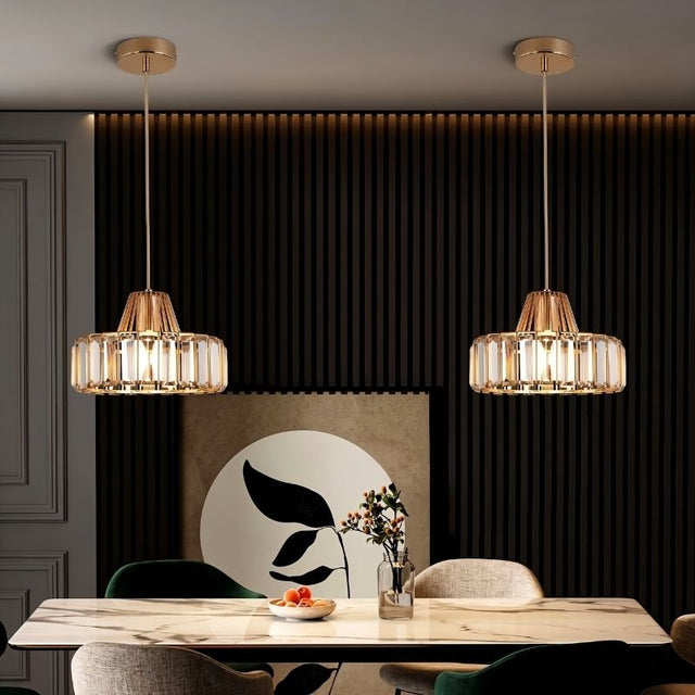  "Two modern crystal pendant lights with gold frames, hanging above a dining table with a marble top and artwork."