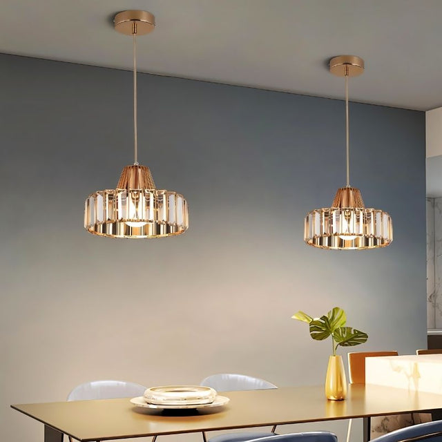 "Two modern crystal pendant lights with gold frames, hanging above a dining table with a plant and place settings."