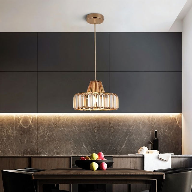 "A modern crystal pendant light with a gold frame, hanging above a kitchen island with a dark countertop and fruit bowl."