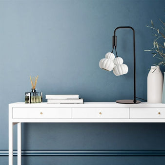 "A modern, two-headed table lamp with metal and ceramic accents in a contemporary home office setting with a white desk and blue wall."