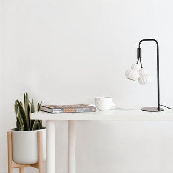 "A modern table lamp with two adjustable heads, combining metal and ceramic elements, illuminating a contemporary home office with a white desk and a plant."