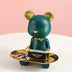 Teal and gold bear figurine holding a luxury handbag and accessories on a white surface with pink background.