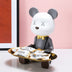White bear figurine with closed eyes and gold bow tie holding a gold tray of packaged items against a pink background.