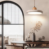 "Modern pendant light with a circular travertine shade and wooden sphere, illuminating a round dining table with decorative items and a large window with blinds."