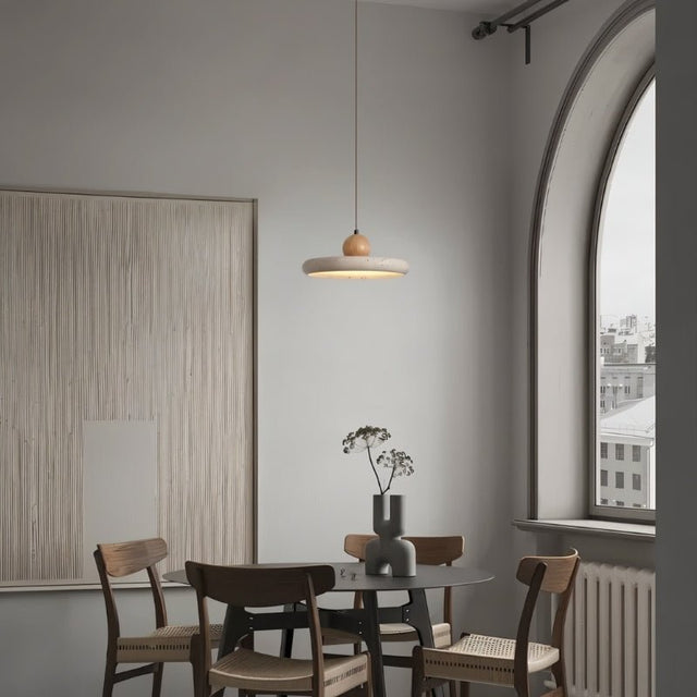 "Modern, minimalist pendant light with a circular travertine shade and wooden sphere, hanging in a dining room with a round table, chairs, artwork, and a large arched window."