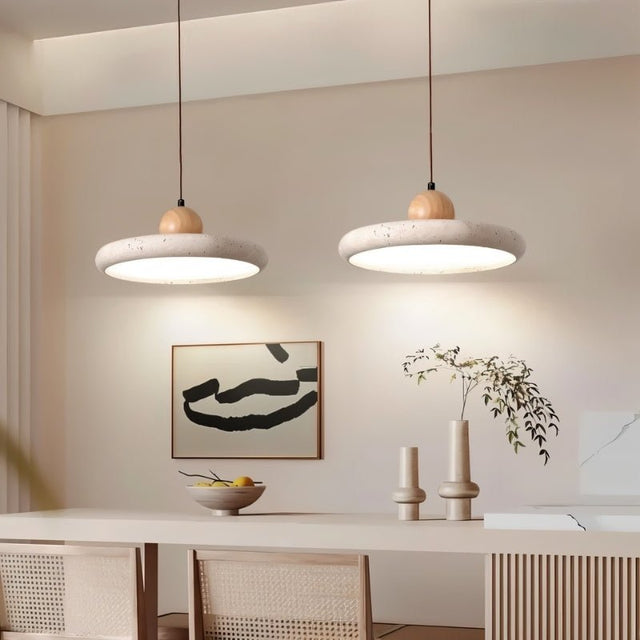 "Pair of modern pendant lights with circular travertine shades and wooden spheres, hanging over a dining table with chairs, artwork, and decorative vases."
