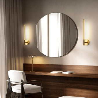 "Two gold Valtie wall lamps with slim, vertical design flanking a round mirror in a bedroom."